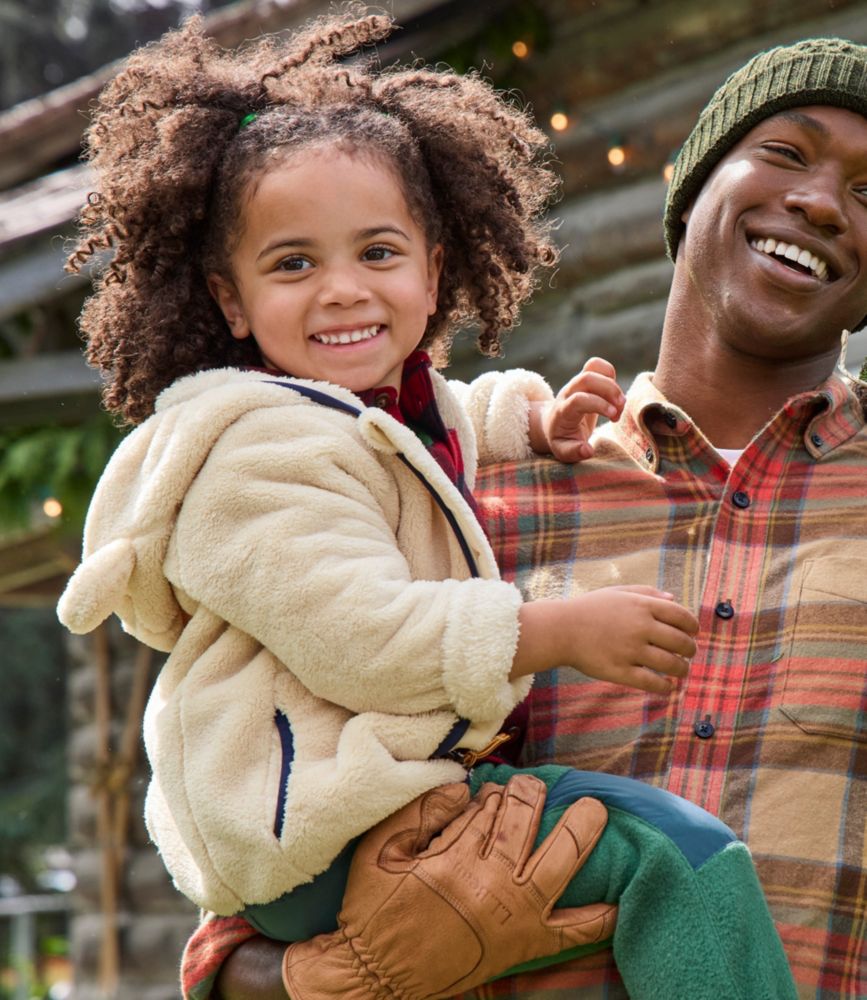 Toddler wearing a natural high-pile fleece jacket with hood, held outdoors by an adult in a plaid shirt.