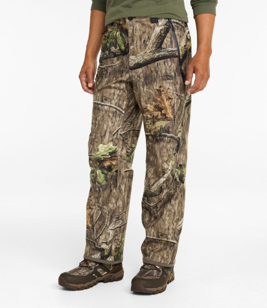 Men’s camo hunting pants with woodland tree and leaf pattern, shown worn with hiking boots against a plain background.
