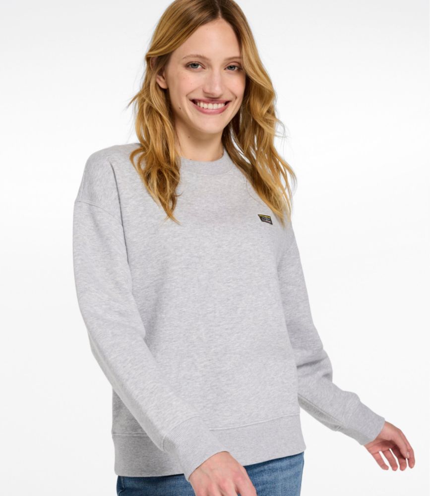 Woman wearing a light gray crewneck sweatshirt with small chest logo and ribbed cuffs, paired with blue jeans.